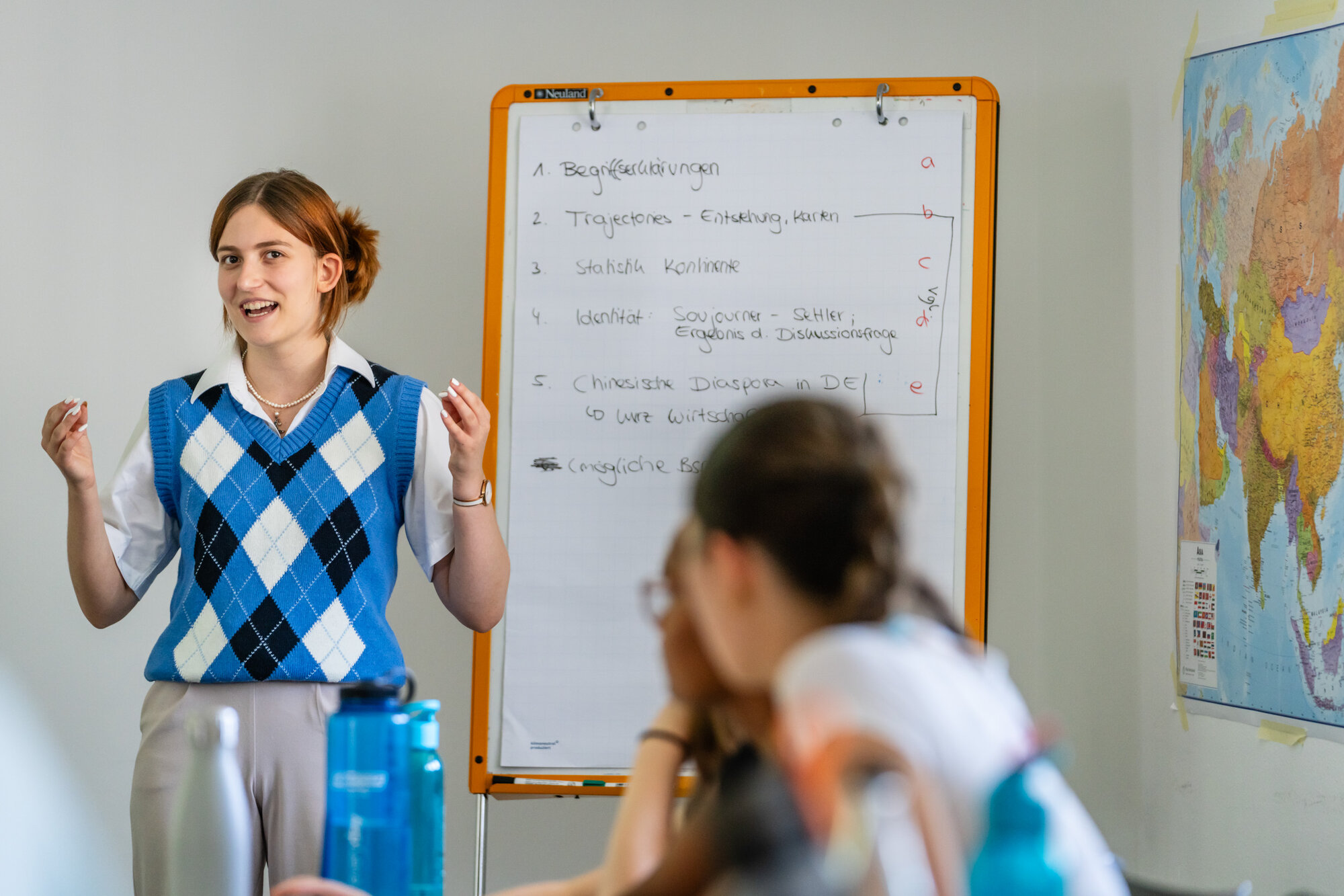 Ein Mädchen im karierten Pullunder steht vor einem Flipchart und gestikuliert. 