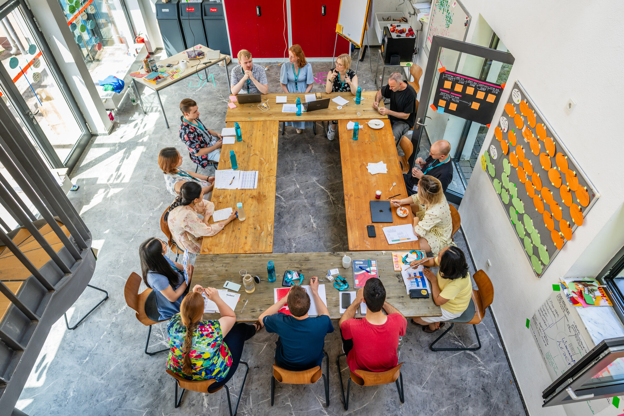 Vierzehn Kursleitungen sitzen um einen großen Tisch. Auf dem Tisch liegen Notizhefte und Stifte. Im Hintergrund ist ein Flipchart zu sehen, und bunte Post-its sind an die Wand geheftet.